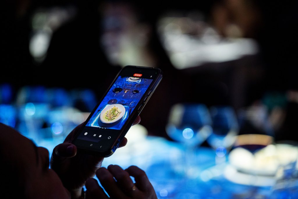 Guest capturing a dish at The Radiant Table on phone with immersive blue lighting