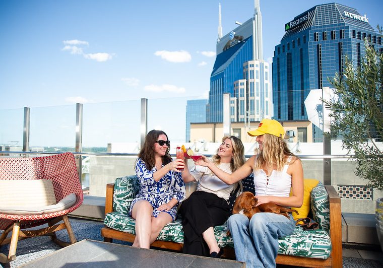 Three women enjoying drinks on a rooftop lounge in Nashville with city skyline in the background.