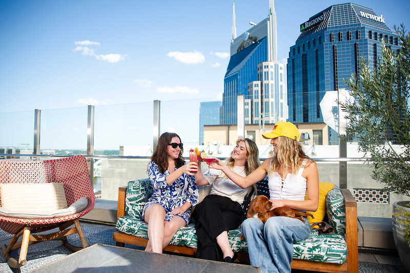 Three women enjoying drinks on a rooftop lounge in Nashville with city skyline in the background.