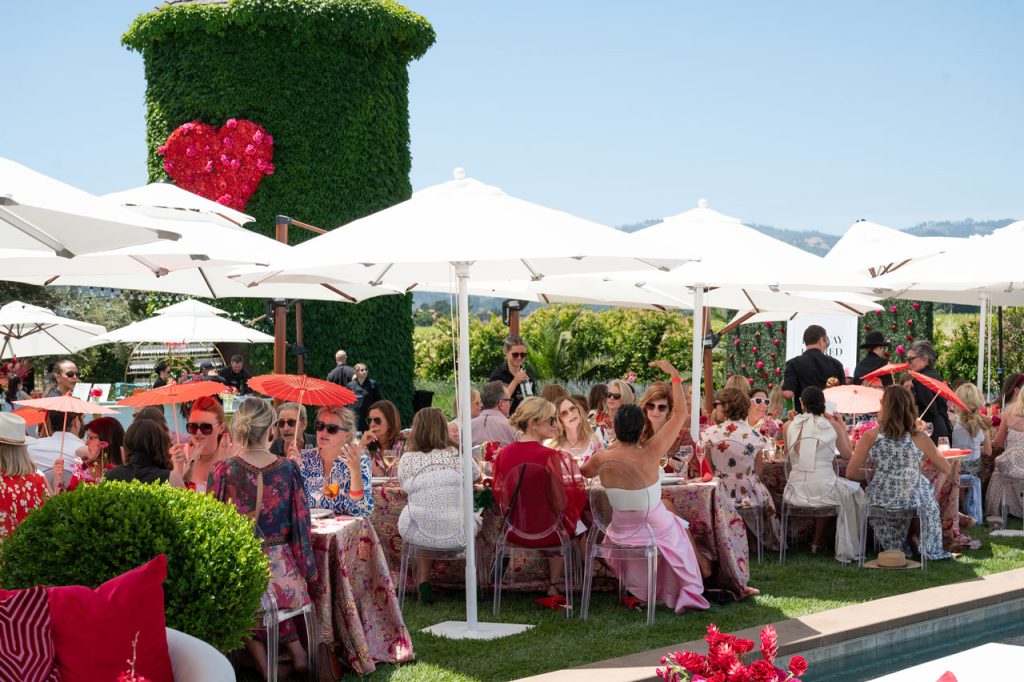Elegant outdoor luncheon at Runway for Red with women in stylish outfits and red parasols gathered under white umbrellas.