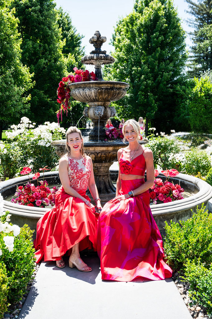 Two women in vibrant red gowns pose by a flower-adorned fountain at the Runway for Red heart health fundraiser in Napa Valley.