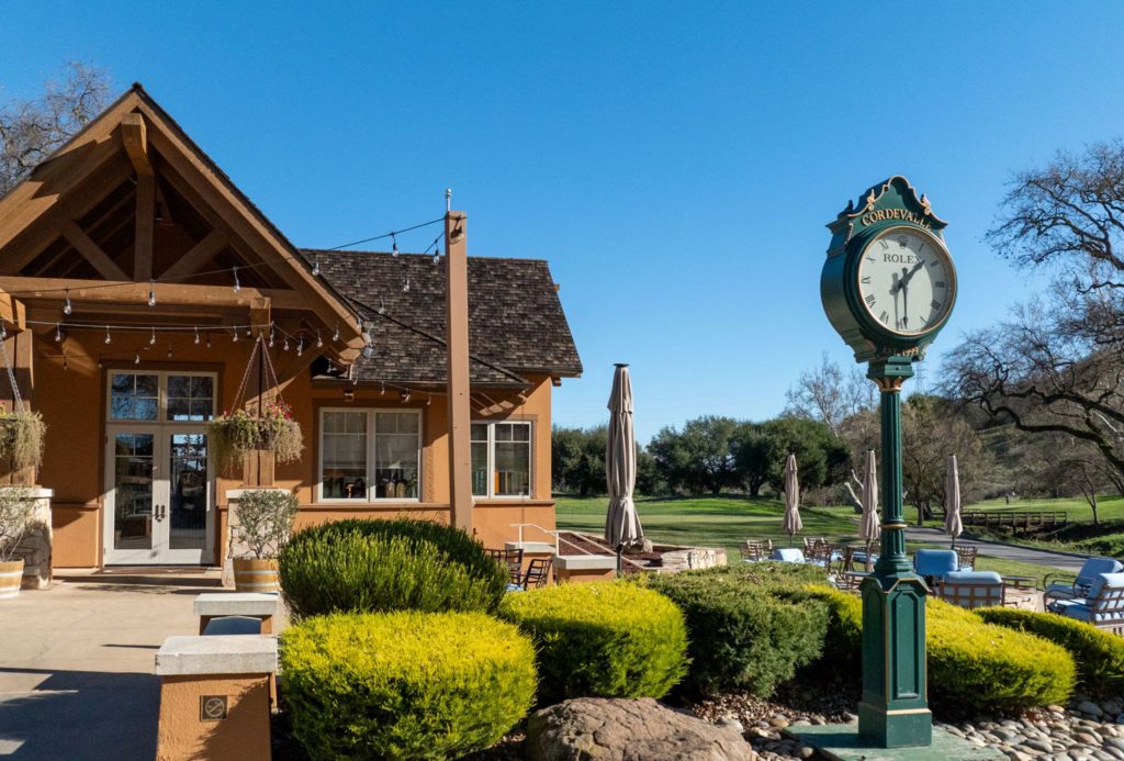 Charming clubhouse patio at CordeValle with vintage Rolex clock and relaxed seating surrounded by lush greenery.
